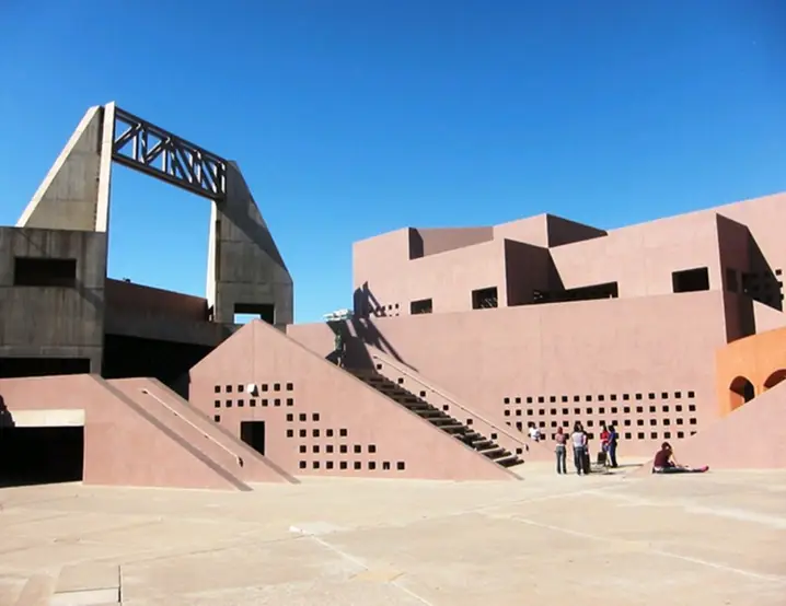 Brutalist concrete architecture with pink geometric buildings, angular ramps, and perforated walls under clear blue sky