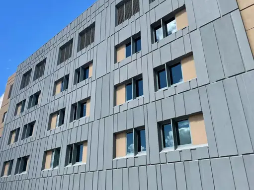 Modern building facade with gray geometric metal panels, recessed windows, and warm interior lighting visible through glass