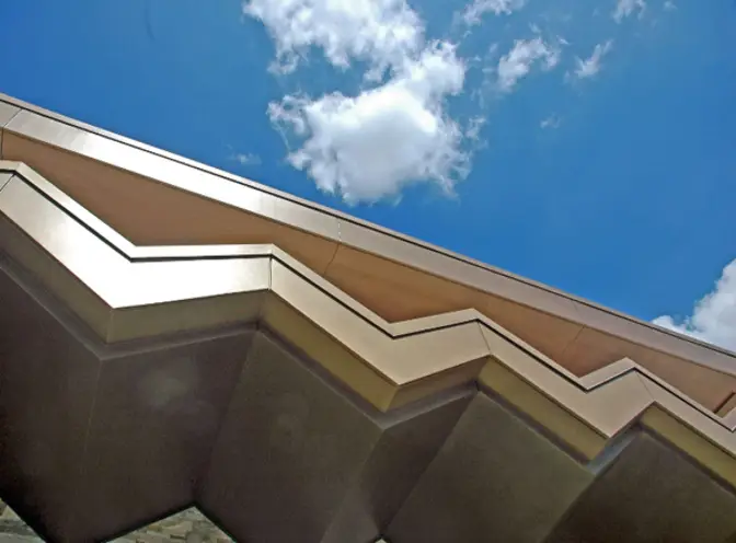 Architectural detail showing zigzag concrete overhang with geometric shadows against bright blue sky and white clouds